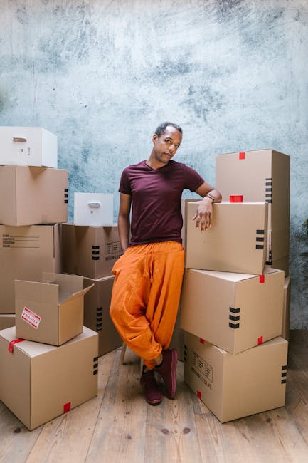 A woman with dark skin and short hair stands in a room with a textured pale blue wall and wooden flooring, surrounded by stacked cardboard boxes of various sizes; some boxes have red and black tape securing them, and a few are open revealing packaging materials or contents. She is wearing an orange pair of trousers and a dark red t-shirt, leaning slightly against a large box with her right arm resting on top of it, while her left hand touches a smaller box. The scene appears to depict a packing or home relocation process, aligning with furniture transport and moving services offered by Man with Van Lower Clapton. A part of a white doorframe is visible on the left, and a van may be nearby for loading, indicating an active loading process for house removals or packing and moving tasks.