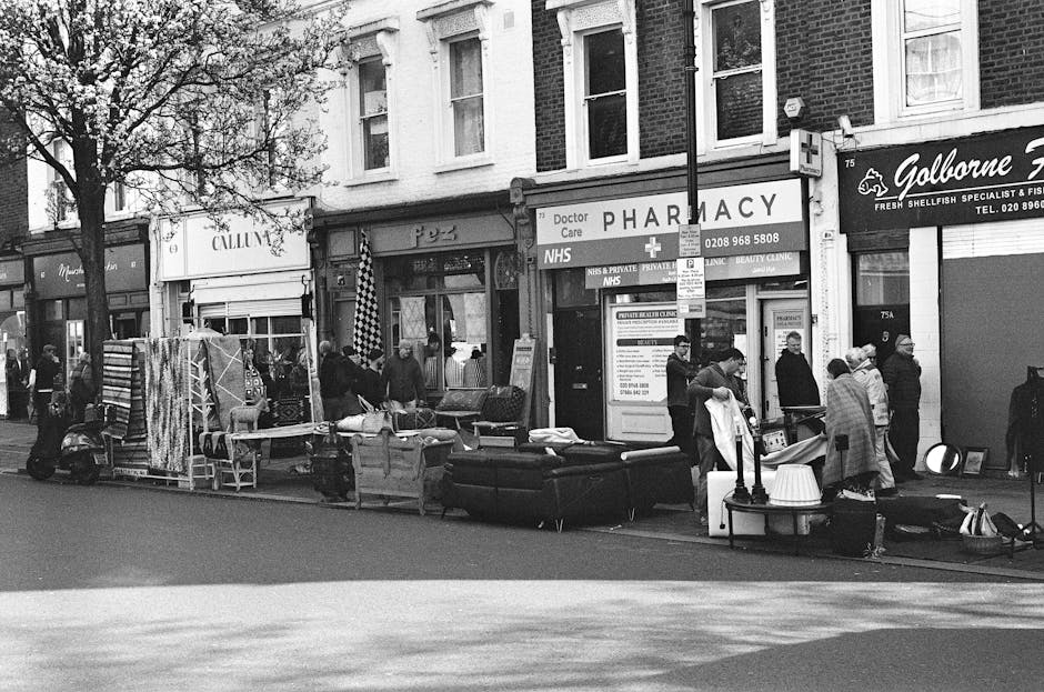 A street scene outside a shopfront with a sign indicating pharmacy services, where a group of people are gathered on the sidewalk. In front of the shop, there are various pieces of furniture including a large black sofa, a wooden table, and a cardboard box. Additionally, there are several wrapped items, possibly furniture or household goods, and a small trolley loaded with belongings. A man in the foreground is handling a large item, possibly for removal or transport, while others stand nearby engaged in conversation or observing. The surroundings include other shopfronts, one selling shellsfish, and a Shop with a sign for Doctor Care, with retail displays and commercial signage visible. The scene appears to be part of a home relocation or furniture transport process, with items arranged closely on the pavement for loading, consistent with moving services provided by companies like Man with Van Lower Clapton.