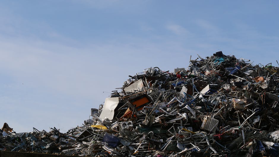 A large pile of metal scrap and discarded household appliances, including broken ovens, washing machines, metal frames, and various electronic waste, stacked on a paved surface outdoors under a partly cloudy sky. The debris consists of twisted and bent materials, with some parts covered in plastic and others exposed as rusted or damaged. In the background, the sky is visible with soft white clouds, indicating an open area near a waste disposal or recycling site. This image highlights waste management and recycling processes related to bulky item removals, which [COMPANY_NAME] handles during home relocation and furniture transport services in the E5 Lower Clapton area.