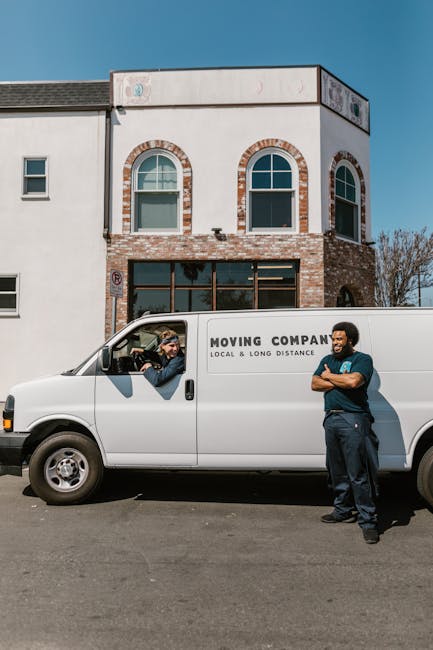 A white moving van parked in front of a two-storey residential building with a brick and white exterior, under a clear blue sky. The van belongs to a local moving company and has the words 'MOVING COMPANY, LOCAL & LONG DISTANCE' printed on the side. Inside the driver's seat, a person is visible handing over a map or document to a technician standing outside the van, who is dressed in dark clothing with arms crossed, smiling. The loading process involves minimal visible objects in the scene, with no furniture or boxes in view, but the context suggests preparations for house relocation or furniture transport, typical of professional removals services as seen in the relevant page about Chatsworth Road moves by Man with Van Lower Clapton.