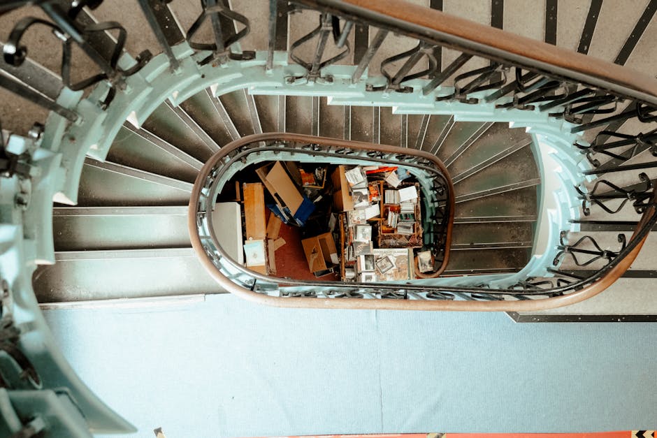 A view from the top of a spiral staircase inside a building, showing books, cardboard boxes, and other miscellaneous items placed on the staircase's landings below. The staircase has a mixture of metal steps, a wooden handrail, and black metal balusters. The items appear to be packed for a home relocation or furniture transport, consistent with a moving or packing and moving process, as seen in house removals services offered by Man with Van Lower Clapton. The corridor at the top of the stairs is carpeted, and the lighting is natural, illuminating the cluttered collection of objects being prepared for loading into a van for transportation, part of a stair solutions move in Clapton Common.