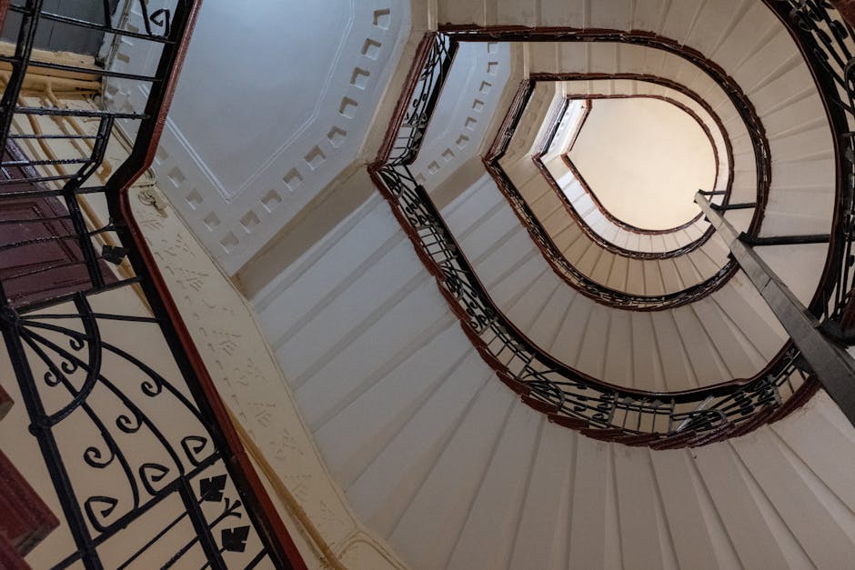 A view from the top of a spiral staircase inside a building, showing books, cardboard boxes, and other miscellaneous items placed on the staircase's landings below. The staircase has a mixture of metal steps, a wooden handrail, and black metal balusters. The items appear to be packed for a home relocation or furniture transport, consistent with a moving or packing and moving process, as seen in house removals services offered by Man with Van Lower Clapton. The corridor at the top of the stairs is carpeted, and the lighting is natural, illuminating the cluttered collection of objects being prepared for loading into a van for transportation, part of a stair solutions move in Clapton Common.
