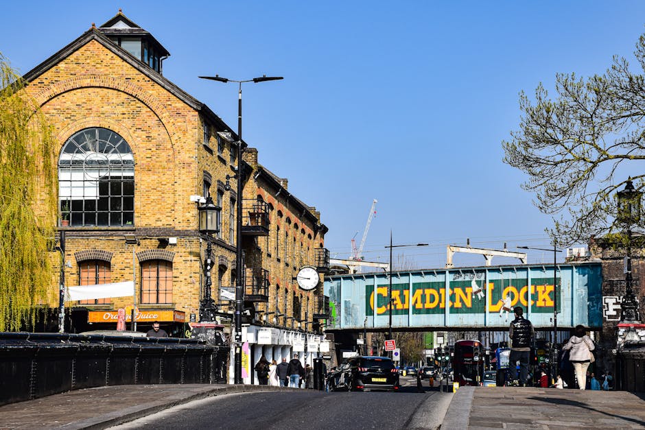 A street scene in Lower Clapton featuring a large brick building with a prominent arched window, a street clock mounted on a pole, and a teal coloured railway bridge with bold yellow and green lettering that reads 'Camden Lock'. Pedestrians are walking along the sidewalk and cars are parked or driving on the road. In the foreground, a black van is visible, suggesting furniture transport or home relocation activities as part of a house removal process. The environment is illuminated with bright daylight, and there are trees with budding leaves on the right side of the image. This scene captures the typical urban setting associated with moving and packing logistics in the context of house removals and transport services in Lower Clapton, as provided by Man with Van Lower Clapton.