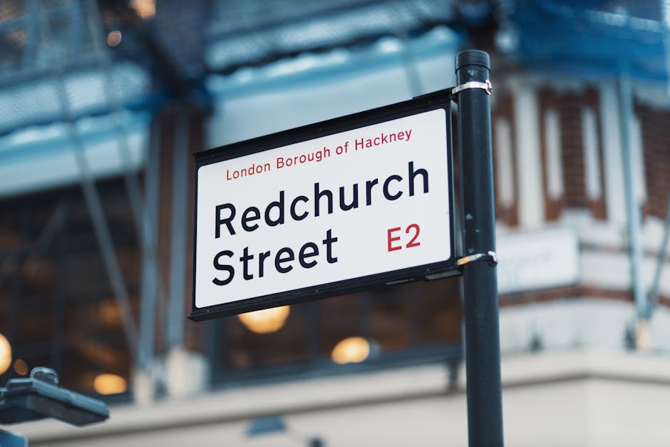 A street sign mounted on a black pole displaying 'Redchurch Street' in bold black letters with the postcode 'E2' in red beneath, indicating a location within the London Borough of Hackney. The sign is positioned at an outdoor setting on a pavement, with a modern building visible in the background, partially obscured by a blue scaffolding structure. The area appears well-lit with natural daylight, and the scene is associated with urban logistics for house removals or furniture transport, as typical for a professional moving service like Man with Van Lower Clapton specializing in relocations within the Hackney area.