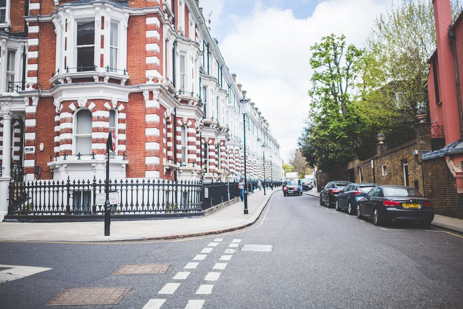 A street scene in Lower Clapton featuring a large brick building with a prominent arched window, a street clock mounted on a pole, and a teal coloured railway bridge with bold yellow and green lettering that reads 'Camden Lock'. Pedestrians are walking along the sidewalk and cars are parked or driving on the road. In the foreground, a black van is visible, suggesting furniture transport or home relocation activities as part of a house removal process. The environment is illuminated with bright daylight, and there are trees with budding leaves on the right side of the image. This scene captures the typical urban setting associated with moving and packing logistics in the context of house removals and transport services in Lower Clapton, as provided by Man with Van Lower Clapton.