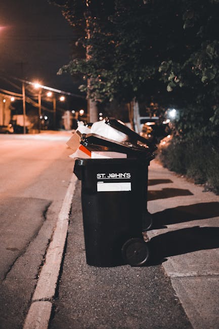 A black wheeled rubbish bin labeled 'St. John's' positioned on the pavement beside a row of dark green bushes and a tree, with a partially open lid revealing various waste items including cardboard boxes, plastic packaging, and paper. The bin is situated next to a residential street at night, illuminated by streetlights casting a warm glow. In the background, a street with parked cars, streetlights, and a pedestrian crossing can be seen, indicating an urban environment. This scene depicts rubbish collection or disposal, relevant to house removals or domestic waste removal services by Man with Van Lower Clapton. The image emphasizes the process of waste clearance in a typical street setting, supporting the content about local rubbish removal rules and logistics involved in home relocation in Lower Clapton.