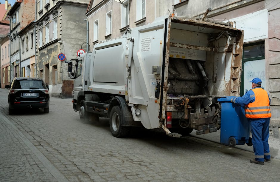 A street scene in Lower Clapton shows a worker from Man with Van Lower Clapton placing a blue rubbish bin into a waste collection truck parked on a cobblestone road. The worker, dressed in a blue uniform and an orange high-visibility vest, is positioned beside the truck's open rear loading hatch, handling the bin with both hands. The waste collection vehicle, which is white and slightly rusted at the rear, is on the street adjacent to a row of multi-story residential buildings with various facades and windows. A black car is parked further down the street, and street signs indicating parking restrictions are visible on the building wall. The environment is lit by natural daylight, and the overall scene reflects rubbish removal as part of home relocation or decluttering processes, consistent with house removals and moving logistics facilitated by Man with Van Lower Clapton.