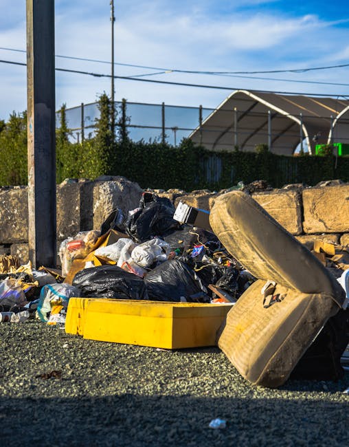 A black wheeled rubbish bin labeled 'St. John's' positioned on the pavement beside a row of dark green bushes and a tree, with a partially open lid revealing various waste items including cardboard boxes, plastic packaging, and paper. The bin is situated next to a residential street at night, illuminated by streetlights casting a warm glow. In the background, a street with parked cars, streetlights, and a pedestrian crossing can be seen, indicating an urban environment. This scene depicts rubbish collection or disposal, relevant to house removals or domestic waste removal services by Man with Van Lower Clapton. The image emphasizes the process of waste clearance in a typical street setting, supporting the content about local rubbish removal rules and logistics involved in home relocation in Lower Clapton.