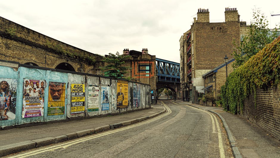 A narrow, curving street in Lower Clapton with a double yellow line, leading toward a blue metal bridge and brick buildings, during daytime with overcast sky. On the left side, a weathered metal fence is covered with colorful posters advertising local events, with some posters torn or faded. Behind the fence, parts of a brick wall and some greenery are visible. The pavement on the right side is bordered by a brick wall partially covered with dense green ivy or hedge. The street appears to be used for residential or local access, evidenced by the quiet surroundings and lack of traffic. This scene is relevant to house removals and moving services, as it depicts the typical environment where furniture transport and home relocation logistics, such as loading and unloading during a move, take place, highlighting the importance of careful planning around urban street layouts. Exterior lighting ensures good visibility of all objects in the image, which could relate to the work of [COMPANY_NAME] in facilitating smooth move routes in the Lower Clapton area.