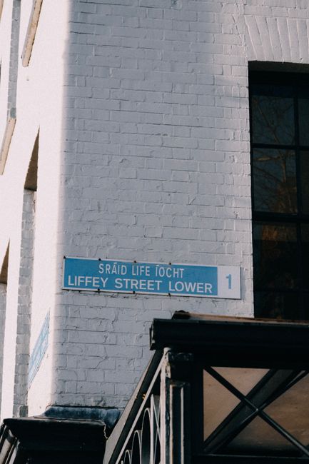 A white brick building with a blue street sign reading 'Liffey Street Lower' and the Welsh translation above, mounted on the exterior wall. The image captures part of a staircase with black metal railings descending towards the pavement, where several cardboard boxes and wrapped furniture pieces are being prepared for a home relocation. Inside the property, visible through a window, are additional packed items and furniture, indicating an ongoing packing and moving process. A black van is partially visible in the foreground, positioned on the street for loading or unloading goods as part of the furniture transport and removals service provided by Man with Van Lower Clapton. The environment is well-lit, suggesting daytime, with the setting focusing on the preparation for a professional moving service involving careful packing, loading, and transportation of household contents along narrow streets and tight turns typical of the area.