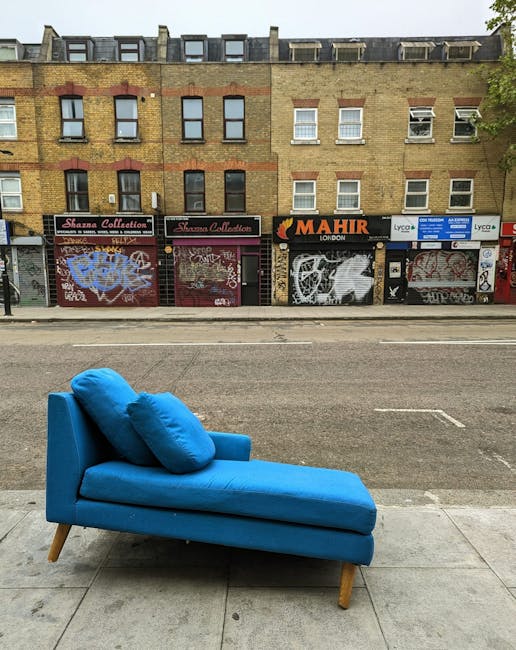 A bright blue upholstered chaise lounge with matching cushions positioned on a sidewalk in front of a row of shopfronts on a city street. The furniture features wooden legs and appears to be in the process of being moved or temporarily placed outdoors. Behind it, the shops have closed metal shutters covered in graffiti, with signs for businesses such as 'Mahir London' and 'Lyca'. The street surface includes a double yellow line, and the scene appears to be during daylight with natural lighting. This setting suggests a home relocation or furniture transport scenario handled by Man with Van Lower Clapton, with focus on the logistics of moving household furniture along narrow streets and tight turns.