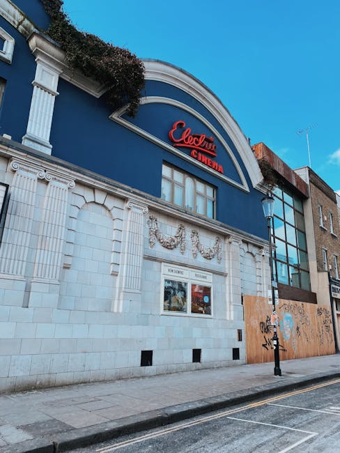 The exterior of a building featuring the Electric Cinema in Lower Clapton with a large blue facade, classical architectural details in white stone including decorative friezes and columns, and a prominent red sign displaying 'Electric Cinema'. The building has a curved arch at the top of the facade and white window frames below the sign. To the right, part of an adjacent structure with modern glass windows is visible, alongside a black lamppost and some graffiti on a wooden barrier. The scene is set on a pavement beside a quiet street with an overcast sky. This image represents the kind of urban environment where house removals or furniture transport services by Man with Van Lower Clapton might coordinate pickup or delivery as part of residential moving logistics.