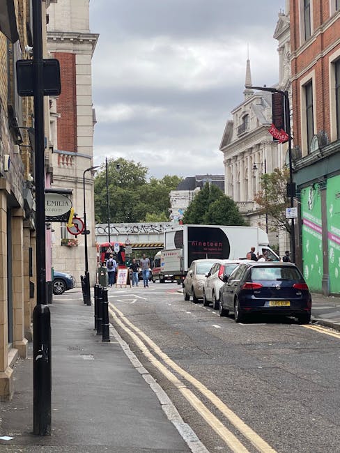 A narrow street in Lower Clapton during a home relocation process, featuring the loading area for a house move. The scene includes several parked cars along the curb, a white moving van positioned towards the background with the rear doors open, and visible cardboard boxes and wrapped furniture inside the vehicle. The pavement in front of the van has a few people assisting with the loading and unloading of household items using trolleys and straps. Building facades line both sides of the street, with a mix of brick and stucco exteriors, some storefront signs, and a green construction barrier visible on the right. Overhead, there are traffic signs, lamp posts, and an overpass running above the street. The sky is overcast with grey clouds, indicating a typical rainy day suitable for interior packing and furniture transport, as part of the house removal services provided by Man with Van Lower Clapton.